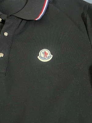 Svart Moncler pikétröja - Klassisk svart pikétröja från Moncler med vit och röd rand på krage och ärmslut. Tröjan har korta ärmar, knappar framtill och Moncler-logga broderad på bröstet. Perfekt för en clean och stilren look.