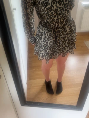 Leopardmönstrad klänning - Säljer en kort klänning med leopardmönster i beige och svart. Klänningen har långärmade, lätt transparenta ärmar, markerad midja och volangdetaljer vid kjolen. Öppen rygg med knapp i nacken ger en snygg detalj.