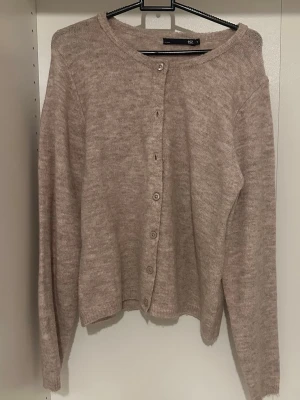 Beige stickad kofta med knappar - Mysig beige stickad kofta med rund halsringning och långa ärmar. Koftan har en enkel design med knappar framtill och ribbade muddar vid ärmslut och nederkant. Perfekt att slänga på sig när det är lite kyligt.