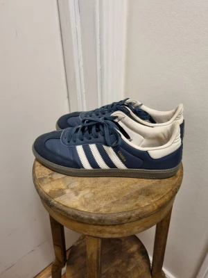 Adidas Samba blå och vita sneakers - Klassiska Adidas Samba sneakers i blått med vita detaljer. Ovandelen är i läder och mocka. Strl 43,5 men passar även 43 Släppt lite på ena skon som förmodligen går att limma och något litet hål under båda. 