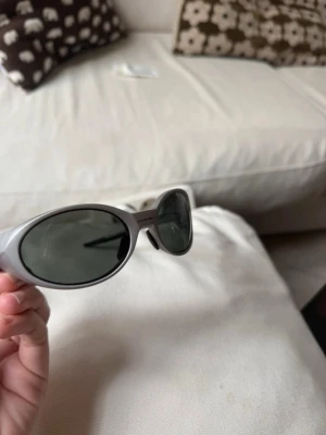 Silvriga ovala solglasögon Oakley - Säljer ett par riktigt snygga solglasögon från Oakley med oval form och silvriga bågar. Glasen är mörka och bågarna har en sportig vibe. Helt nya. Box och fodral tillkommer