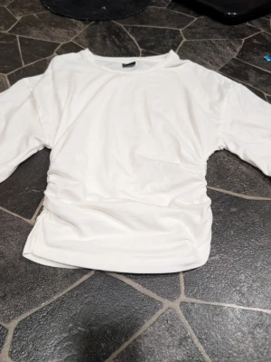 Vit topp med rynkade sidor stl xs - Säljer en vit topp med rund hals och rynkade detaljer på sidorna. Toppen har korta, lite vidare ärmar och är gjord i ett mjukt material som känns skönt mot huden. Perfekt att styla med jeans eller kjol för en clean look.