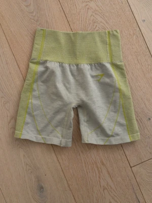 Ljusgråa högmidjade träningsshorts - Säljer ett par ljusgråa högmidjade träningsshorts med ribbad midja och detaljer i gult. Shortsen har en tight passform och är tillverkade i ett stretchigt syntetmaterial som passar perfekt för träning.