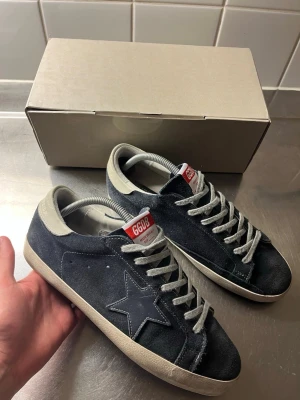 Mörkblåa Golden goose  - Snygga svarta sneakers från Golden Goose med ikonisk stjärndetalj på sidan. Skorna har grå mocka, vita sulor och grå skosnören. Märkets röda och vita GGDB-logga syns på plösen. Perfekta för dig som gillar streetstyle och unika detaljer.