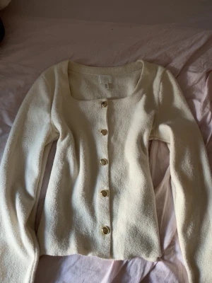 Krämvit kofta från H&M med guldknappar - Snygg krämvit kofta från H&M med långa ärmar och fyrkantig ringning. Koftan har dekorativa guldknappar framtill och är gjord i ett mjukt, strukturerat material som känns lyxigt. Perfekt att styla över ett linne eller t-shirt.