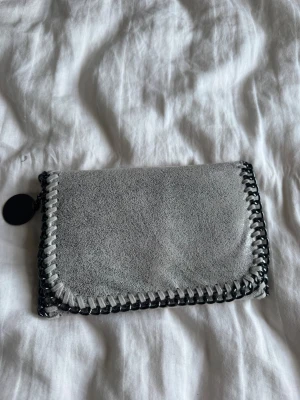 Grå mockaplånbok med kedjedetaljer - Snygg grå plånbok i mocka med coola kedjedetaljer runt kanterna. Plånboken har en rund svart dragkedjeberlock och en mjuk, rektangulär form. Perfekt för dig som gillar stilrena accessoarer med en twist.