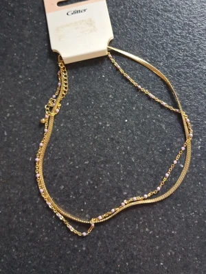 Guldigt halsband  - Från Glitter. Små lila pärlor på andra kedjan. Aldrig använd 