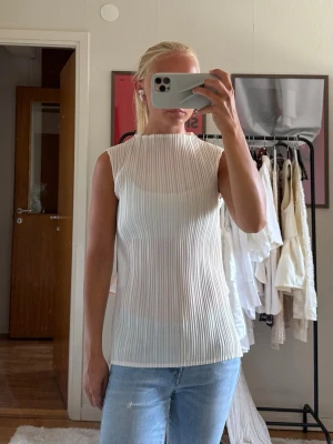 Vit plisserad ärmlös topp - Säljer en stilren vit plisserad topp med hög hals och ärmlös design. Toppen har en lätt transparent känsla och rak passform som ger en modern look. 