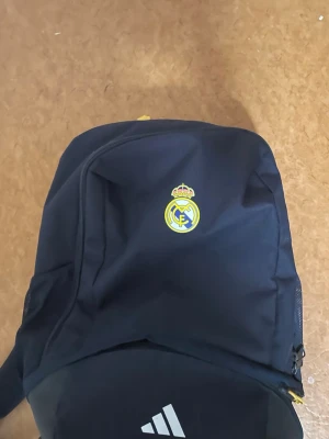 Svart Real Madrid ryggsäck - Snygg svart ryggsäck med Real Madrid-logga på framsidan och vit Adidas-logga nedtill. Ryggsäcken har dragkedja och flera fack, perfekt för skolan eller träningen. Tillverkad i slitstarkt material och har justerbara axelband.