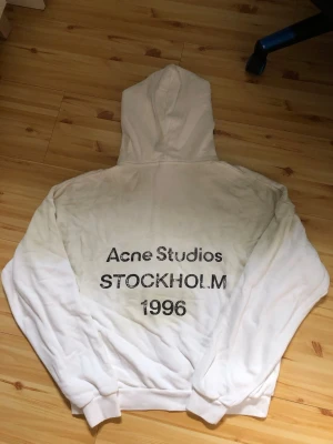 Vit hoodie Acne Studios StockholmÖPPEN FÖR BUD - Säljer en vit hoodie från Acne Studios med trycket 'Acne Studios STOCKHOLM 1996' på ryggen. Hoodien har en klassisk huva och ribbade muddar. Perfekt för dig som gillar streetwear och skandinavisk design.