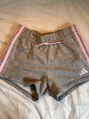 Grå shorts med rosa detaljer Adidas - Gråa shorts från Adidas med ljusrosa ränder längs sidorna och matchande snörning i midjan. Adidas-logga i rosa på ena benet. I ålder 13/14.