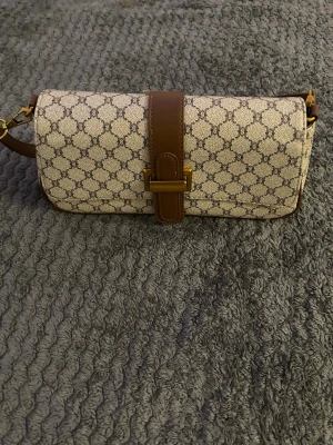 Beige axelremsväska med monogram - Snygg beige handväska med brunt läderband och axelrem. Väskan har ett klassiskt monogrammönster i brunt och guldiga metalldetaljer. Avlång och kompakt modell som stängs med ett spänne framtill. Perfekt för dig som gillar stilrena accessoarer.