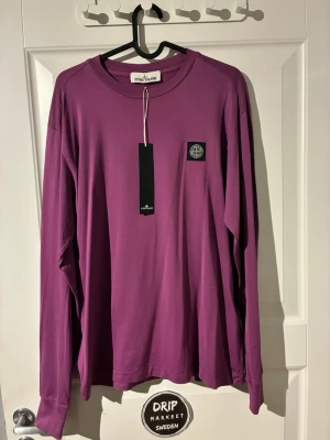 Lila långärmad tröja från Stone Island - Säljer en stilren lila långärmad topp från Stone Island i storlek medium med klassisk rund hals och ikonisk patch på bröstet. Tillverkad i mjuk bomull som känns skön mot huden. Perfekt för dig som vill ha en clean och trendig look.