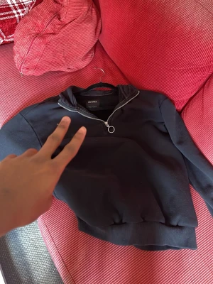 Svart hoodie med halv dragkedja - Säljer en svart hoodie med halv dragkedja och ringdetalj vid öppningen. Hoodien har ribbade muddar vid ärmslut och nederkant samt en hög krage. Perfekt för en chill och avslappnad stil.