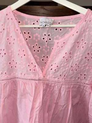 Rosa broderad klänning från Noella - Söt ljusrosa klänning från Noella med broderade blommor och v-ringning. Klänningen har volangdetaljer och är tillverkad i ett lätt bomullstyg som passar perfekt till varmare dagar. 
