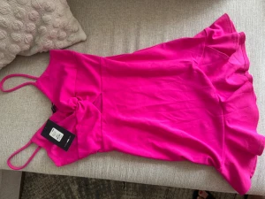 Kort rosa klänning med volang - Snygg kort klänning i starkt rosa med tunna axelband och volangkant nedtill. Klänningen har en twistad detalj framtill och är figurnära i modellen. Perfekt för dig som vill sticka ut med färg och enkel design.