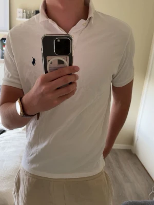 Vit pikétröja från Ralph Lauren - Klassisk vit pikétröja från Ralph Lauren med blå logga på bröstet. Tröjan har korta ärmar, krage och knappar vid halsen. Modellen är custom slim fit och passar perfekt till en avslappnad men stilren look. Storlek L