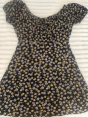 Svart blommig kortklänning - Söt svart kortklänning med små vita och gula blommor. Klänningen har korta ärmar och en lätt utställd passform. Perfekt för varma dagar och har en ungdomlig, fräsch look med sitt blommiga mönster.
