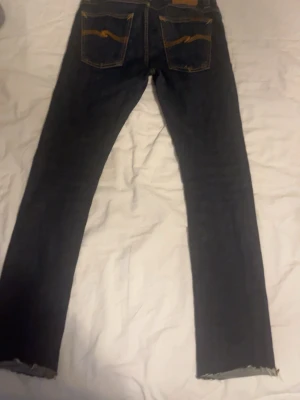Mörkblå jeans med rå kant - Säljer ett par mörkblå jeans med smal passform och rå, fransig kant vid bensluten. Jeansen har klassiska bakfickor med kontrastsömmar och är perfekta för en avslappnad och trendig look.