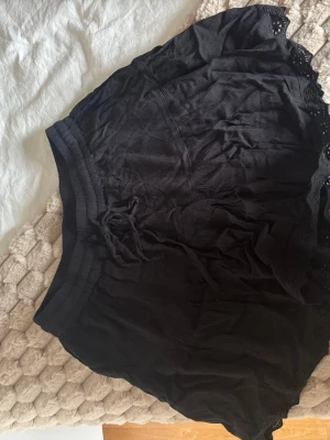 Svart kortkjol med spetsdetaljer - Säljer en svart kortkjol med bred resår i midjan och knytband. Kjolen har fina spetsdetaljer längs nederkanten och är gjord i ett lätt, luftigt material som passar perfekt till varma dagar.