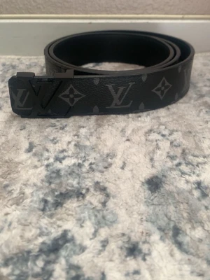 Svart Louis Vuitton bälte i skinn - Snyggt svart bälte från Louis Vuitton i skinn med klassiskt monogrammönster och mattsvart LV-spänne. Perfekt accessoar för att lyfta din outfit med en exklusiv touch.