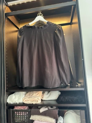 Svart blus med puffärm - Stilren svart blus med rundad halsringning och diskreta puffärmar. Blusen har lång ärm med lätt rynk vid axlarna och muddar vid ärmslut. Materialet ser ut att vara mjukt och har en lätt glansig finish, vilket ger en elegant känsla.