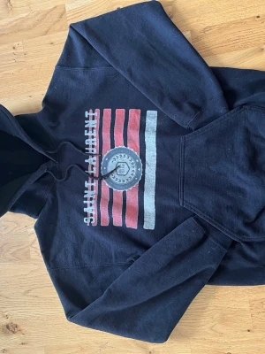 Svart Saint Laurent hoodie med tryck - Säljer en svart hoodie från Saint Laurent med huva och dragsko. Framsidan har ett stort tryck med röda och vita ränder samt ett runt emblem i mitten. Hoodien har en klassisk känguruficka och är gjord i mjukt bomullsmaterial. Perfekt för en avslappnad och cool stil. Såklart äkta, öppen för byten och pris ej hugget i sten!