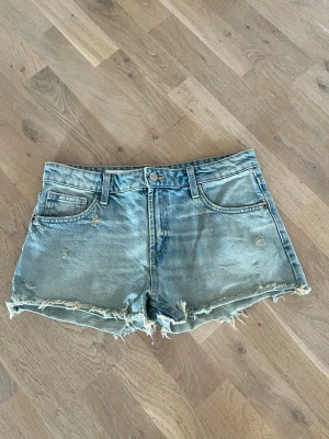 Ljusblå jeansshorts från Zara - Säljer ett par ljusblå jeansshorts från Zara i klassisk denim med slitna detaljer och fransig kant nedtill. Shortsen har fem fickor, bälteshällor och knappgylf. Perfekta för varma dagar och en avslappnad stil.