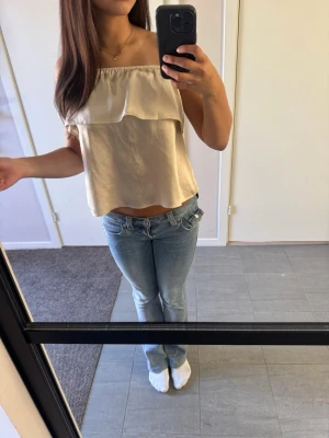 Beige axelbandslös topp med volang - Säljer en beige topp med volangdetalj upptill. Toppen är croppad och har en luftig passform, perfekt för varma dagar. Tillverkad i ett mjukt och lätt material som ger en skön känsla. Men kan ha den på två olika sätt som ni ser på bilden. Storlek 146/152 men känns större.
