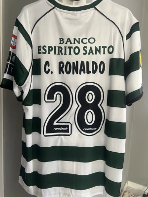 Sporting Lissabon tröja C. Ronaldo #28 - Klassisk fotbollströja från Sporting Lissabon med gröna och vita ränder, C. Ronaldo och nummer 28 på ryggen. Märkt med Reebok och klubbmärke på bröstet. Korta ärmar och flera sponsorloggor. Perfekt för fans och samlare!