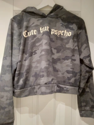 Grön camo / camouflage hoodie, Cute but psycho, Strlk Liten XL, som S-M - Säljer en grönmelerad hoodie med camouflagemönster och texten 'Cute but psycho' i gotisk stil på bröstet. Hoodien har huva och är croppad i modellen. Perfekt för dig som gillar streetwear och vill sticka ut. Strlk XL men är mer som S-M. 