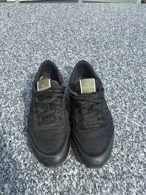 Svarta Valentino sneakers - Snygga svarta rockrunners från Valentino med klassisk låg profil. Skorna har ovandel i mesh och mocka, samt robust gummisula med rutmönster. Tidlös design med svarta skosnören och diskret Valentino-logga på plösen. Storlek 40 men passar även 41