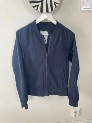 Mörkblå jacka från WearColour - Snygg mörkblå jacka från WearColour med minimalistisk design. Jackan har dragkedja framtill, två sidofickor och ribbade muddar vid ärmslut och nederkant. Tillverkad i lätt och slitstarkt material, perfekt för kalla vinterdagar.
