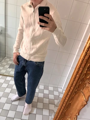 Beige ziptröja från Bläck - Snygg beige ziptröja från Bläck med hög krage och hel dragkedja framtill. Tröjan är i mjukt material och har ribbade muddar vid ärmslut och nederkant. Perfekt för en clean och stilren look.