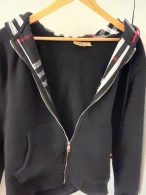 Burberry kofta  - Snygg svart hoodie med dragkedja och fickor framtill. Huvan har ett rutigt mönster i svart, vitt och rött på insidan som ger en unik touch. Tillverkad i mjukt material, perfekt för en chill och avslappnad stil. Broderad logga på bröstet.