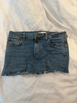 Jeanskjol från Zara - Kort jeanskjol i klassisk blå denim med fem fickor och bälteshällor. Kjolen har en rå, fransig kant nedtill för en avslappnad och trendig look. Perfekt för dig som gillar en edgy stil. Materialet är denim i bomull.