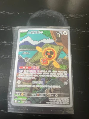 Pokémonkort med färgglad illustration - Säljer ett Pokémonkort med en färgglad illustration av en orange och gul karaktär mot en bakgrund med berg och sjö. Kortet är på koreanska och ligger i ett plastfodral. Perfekt för samlare eller Pokémonfans!
