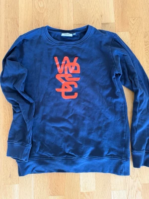 Blå sweatshirt från WESC med tryck - Mörkblå sweatshirt från WESC med rund hals och lång ärm. Framsidan har ett stort rött grafiskt tryck med WESC-loggan. Tröjan är i mjukt bomullsmaterial och har ribbade muddar vid ärmslut och nederkant.