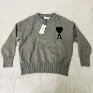 Grå stickad tröja från Ami -  Grå stickad tröja från Ami med svart hjärta och bokstaven A broderat på bröstet. Tröjan har rund halsringning, ribbstickade muddar och är tillverkad i mjukt material. Längd från axelhöjd till midja: 60cm. Pris kan diskuteras😊