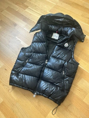 Svart dunväst från Moncler - Snygg svart dunväst från Moncler med glansig finish, avtagbar huva och två dragkedjefickor framtill. Västen har Moncler-logga på bröstet och justerbar nederkant med dragsko. Perfekt för lager på lager och streetwear.