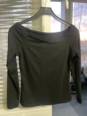 Svart långärmad offshoulder topp - En stilren svart långärmad topp med offshoulder-skärning. Toppen har en smal passform och är tillverkad i mjuk bomull, perfekt för en trendig och enkel look. Passar till många olika outfits och är ett måste i garderoben.