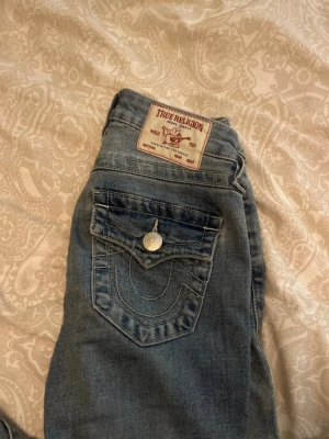 True Religion jeans - Säljer ett par blå bootcut jeans från True Religion (Becca) med klassisk kontrastsöm och ficklock bak. Jeansen har en ljus tvätt och är tillverkade i denim med snyggt slitna detaljer. Perfekta för dig som gillar en avslappnad men trendig stil. Storlek w23, inga defekter förutom lite slitage vid fötterna (sista bilden) . Öppen för flesta prisförslag 