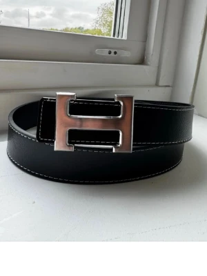 Svart bälte H-spänne - Snyggt svart bälte i skinn med en stor, silverfärgad H-formad metallspänne. Bältet har vit kontrastsöm längs kanterna och en stilren, klassisk look som passar till många outfits.                  ❗️PRIS KAN DISKUTERAS ❗️