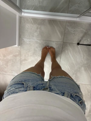 Ljusblå jeansshorts med råa kanter - Säljer ett par ljusblå jeansshorts med slitna detaljer och råa kanter. Shortsen har klassisk femficksdesign, bälteshällor och knappgylf. Perfekta för varma dagar och ger en avslappnad vibe.