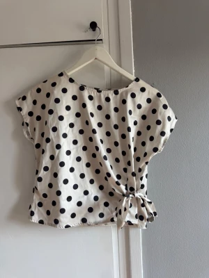 Prickig blus med knyt i midjan - Snygg vit blus med svarta prickar och trendig knytning i midjan. Blusen har korta ärmar och en avslappnad passform, perfekt för dig som gillar en lekfull och stilren look. Tillverkad i ett mjukt och lätt material som känns skönt mot huden.