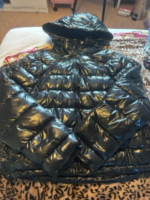 Svart glansig pufferjacka med huva - Säljer en svart, glansig pufferjacka med stor huva och dragkedja framtill. Jackan har breda, vadderade kanaler och elastisk dragsko nedtill för extra värme. Perfekt för kalla vinterdagar och riktigt snygg streetstil.