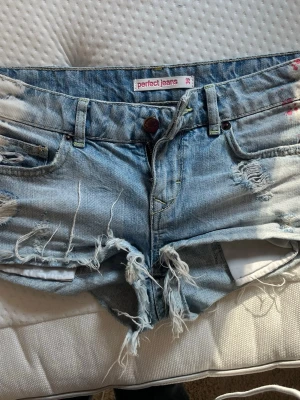 Ljusblå slitna jeansshorts - Säljer ett par ljusblå jeansshorts med slitna detaljer och cool detalj på sidorna. Perfekta för en avslappnad och trendig look.