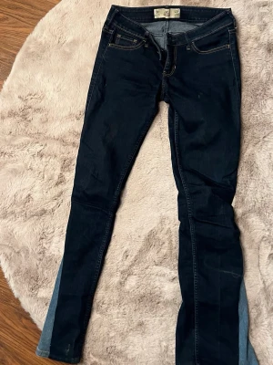 Mörkblå skinny jeans med kontrastpaneler - Säljer ett par mörkblå skinny jeans med ljusblå kontrastpaneler längst ner på benen. Jeansen har klassisk femficksdesign, dekorativa sömmar på bakfickorna och smal passform. Materialet är stretchigt denim för extra komfort.