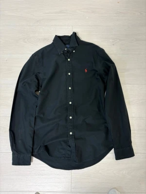 Svart skjorta från Polo Ralph Lauren - Snygg svart skjorta från Polo Ralph Lauren i slim fit-modell. Skjortan har långa ärmar, klassisk krage, knäppning framtill och en röd broderad logga på bröstet. Perfekt för en stilren look.