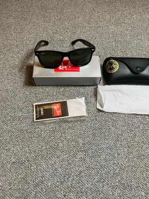Svarta Wayfarer solglasögon Ray-Ban - Klassiska svarta Wayfarer solglasögon från Ray-Ban med gröntonade glas. Snygg och tidlös design med Ray-Ban-logga på linsen. Kommer med originalfodral, putsduk och box. Perfekta för soliga dagar och ger en cool vibe.